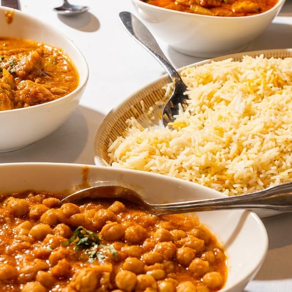 Bowls of chicken curry, chickpea curry, and a platter of basmati rice on a white tablecloth.