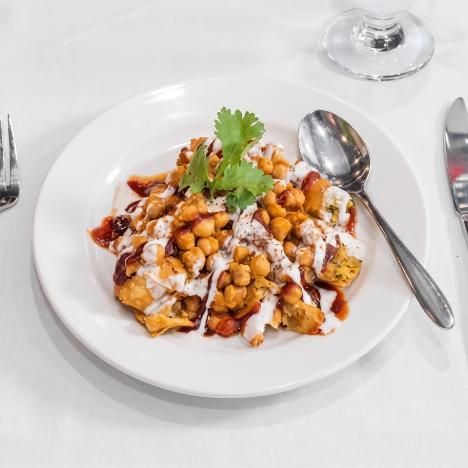 Chickpea chaat with crispy pieces, red and white sauces, and cilantro served on a white plate with silverware.