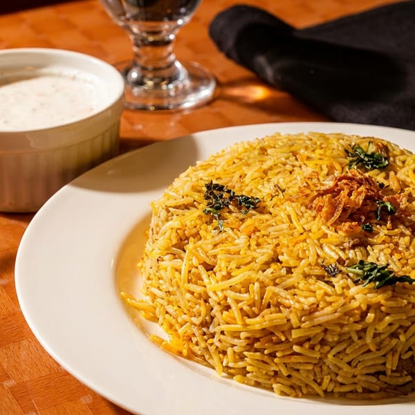 A plate of golden biryani rice with fried onions and herbs, served with a side of raita.