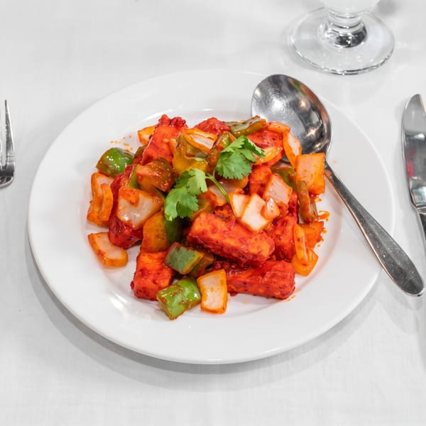 Vibrant red paneer/tofu dish with green peppers and onions, garnished with cilantro, on a white table setting.