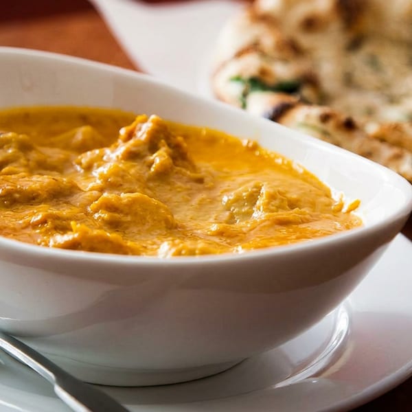 A white bowl of creamy orange curry with a fork, and naan bread in the background.