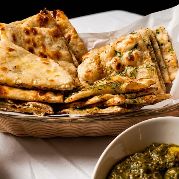 A basket of golden-brown naan bread, some plain and some with herbs, next to bowls of curry.
