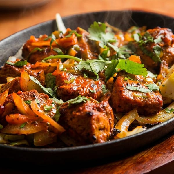 Sizzling chicken tikka with onions, peppers, cilantro, and lemon wedges in a black cast iron pan.